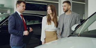友好的汽车销售员正在与自信的年轻人告诉他的新车型，而漂亮的女人是站在她的丈夫旁边，抱着他的手臂和微笑。