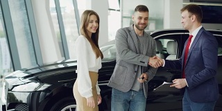 英俊的小伙子正在汽车经销店签购销协议，从推销员那里拿到钥匙链，交给他的妻子，她微笑着亲吻他。