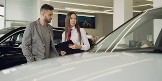 有魅力的女车商拿着文件站在漂亮的车旁边，正在和有兴趣的客户谈论新车型。