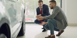 两名兴高采烈的顾客和友好的推销员蹲在一辆昂贵的汽车旁，一边交谈一边打手势讨论汽车型号。购买和销售汽车的概念。