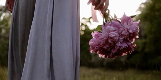 一个穿着鲜艳衣服的女人手持一束粉红色的鲜花走在绿色草地上的背影跟踪录像。阳光。低角度的观点