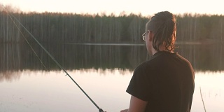 池塘上的渔夫。戴着眼镜梳着辫，穿着t恤用钓竿钓鱼的年轻人。