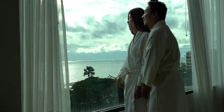 4k视频场景的亚洲夫妇在早上醒来时，睡袍和打开窗户上的窗帘在豪华酒店卧室，生活和休闲概念