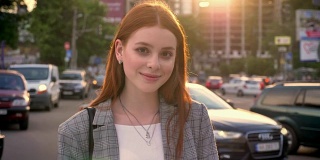 年轻女子的肖像与姜黄色头发看在相机和站在城市街道在日落，微笑，车辆的背景