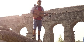 年轻的男子冒险家旅行者站在一个原木树干上弹吉他唱歌在古罗马渡槽遗址在parco degli acquedotti公园在罗马日出慢镜头