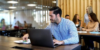 一个帅气的男人在一个共用的办公空间里用他的笔记本电脑工作，在他身后开会的一群人看起来都很高兴