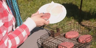 年轻女子把肉排放在烤架上。特写镜头。她穿着粉红色的格子衬衫，梳着蓝色的辫子。