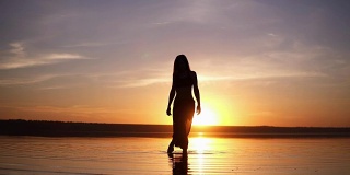 一个美丽的女人的剪影在早晨或傍晚的水面上的前镜头。穿着瑜伽裤的苗条女孩