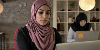 年轻的穆斯林妇女戴着头巾在工作，看着镜头，两个妇女坐在现代办公室的笔记本电脑上打字