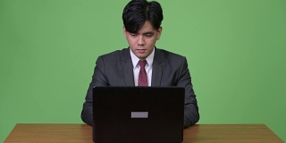 年轻英俊的亚洲商人在绿色背景下用笔记本电脑工作
