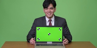 年轻英俊的亚洲商人在绿色背景下展示笔记本电脑
