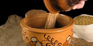 特写镜头，一位老妇人的手从一个木盘子里倒进一个装着麦粒的陶罐里