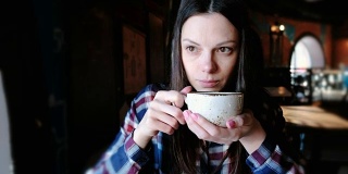 穿着格子衬衫的黑发女人正从一个大杯子里喝茶。疲惫的女人喝杯茶。