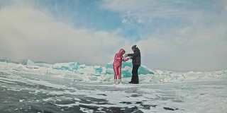年轻的夫妇有乐趣在冬季散步的背景冰冻结的湖。情侣们在一块巨大的蓝色浮冰附近玩得很开心，欢笑、跳跃、亲吻。蜜月。爱情故事。