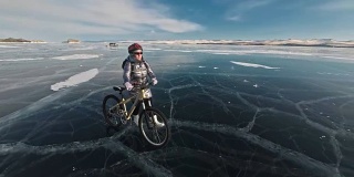 在冰上，一个女人在自行车旁边行走。这个女孩穿着银色的羽绒服，背着背包，戴着头盔。结冰的贝加尔湖的冰。自行车的轮胎上覆盖着特殊的尖钉。