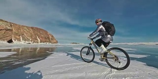 一个女人在冰上骑自行车。这个女孩穿着银色的羽绒服，背着自行车背包，戴着头盔。结冰的贝加尔湖的冰。自行车的轮胎上覆盖着特殊的尖钉。