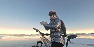 女人在冰上骑自行车。这个女孩穿着银色的羽绒服，背着自行车背包，戴着头盔。女人在用摩擦加热她的手。旅行者正在骑自行车。