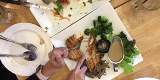 女人手拿刀叉切烤牛排在白色的盘子和食物，另一个是完成在餐馆的木桌上。前视图。视频片段。