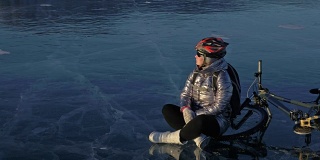 一个女人在冰上骑自行车。这个女孩穿着银色的羽绒服，背着自行车背包，戴着头盔。骑自行车的人停下来休息。她坐在驾驶座上欣赏日落。