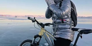 女人在冰上骑自行车。这个女孩穿着银色的羽绒服，背着自行车背包，戴着头盔。女人在用摩擦加热她的手。