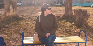 年轻漂亮的黑发女人戴着墨镜环顾四周，等待有人坐在多层城市庭院的长凳上。