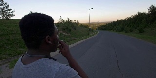 年轻的非洲裔美国人走在空旷的道路上，在日落时吸烟的背影