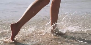 一个年轻女人的腿在海边散步。女子沿着海岸奔跑溅起海水，慢镜头。女人赤脚跑进浅浅的透明海水里。