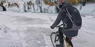 一个人在冰洞附近骑自行车。有冰洞和冰柱的岩石非常漂亮。骑自行车的人穿着灰色羽绒服，背包和头盔。旅行者正在骑自行车。