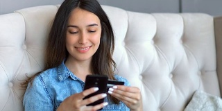 年轻漂亮的女人用信用卡和智能手机在网上购物坐在家里客厅的沙发上