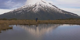 慢动作-旅行者步行横向观看塔拉纳基火山在北岛的新西兰和反射山和登山者在湖中。新西兰被