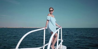 一个年轻美丽的白人女人在游艇上的肖像穿着蓝色的裙子和太阳镜。假期的概念，自由，阳光夏日。背景是蓝色的海洋。steadycam
