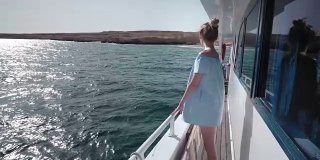 一个年轻美丽的白人女人在一个游艇上的肖像在蓝色的裙子和太阳镜看着一个荒岛。假期的概念，自由，阳光夏日。背景是蓝色的海洋。steadycam