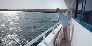 一个年轻美丽的白人女人在游艇上的肖像穿着蓝色的裙子和太阳镜。假期的概念，自由，阳光夏日。背景是蓝色的海洋。steadycam