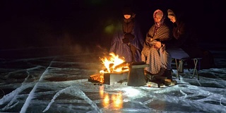 三个旅行者在夜间的冰面上着火。营地在冰上。帐篷紧挨着火。贝加尔湖。附近有一辆车。人们围着篝火取暖，穿着睡袋。