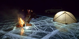 三个旅行者在夜间的冰面上着火。营地在冰上。帐篷紧挨着火。贝加尔湖。附近有一辆车。人们围着篝火取暖，穿着睡袋。