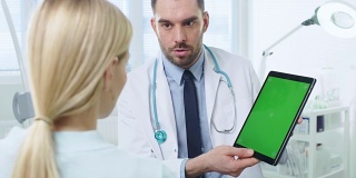 肩膀上方的照片，一位女病人正在与一位专业医生交谈，医生展示了她的绿色屏幕模拟数字平板电脑。