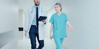 外科医生和女医生一边使用平板电脑一边匆忙地穿过医院走廊，谈论病人的健康。拥有专业人员的现代光明医院。