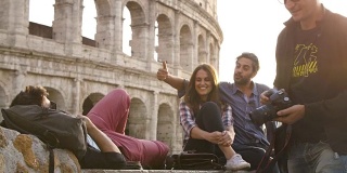 三个年轻的朋友游客坐在罗马的斗牛场前在夕阳下拍照用单反相机背包太阳镜快乐美丽的女孩长发慢镜头