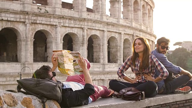 三个年轻的朋友游客坐在罗马斗牛场前在日落阅读地图指导指向用背包太阳镜快乐美丽的女孩长发慢动作