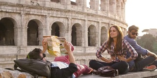 三个年轻的朋友游客坐在罗马斗牛场前在日落阅读地图指导指向用背包太阳镜快乐美丽的女孩长发慢动作