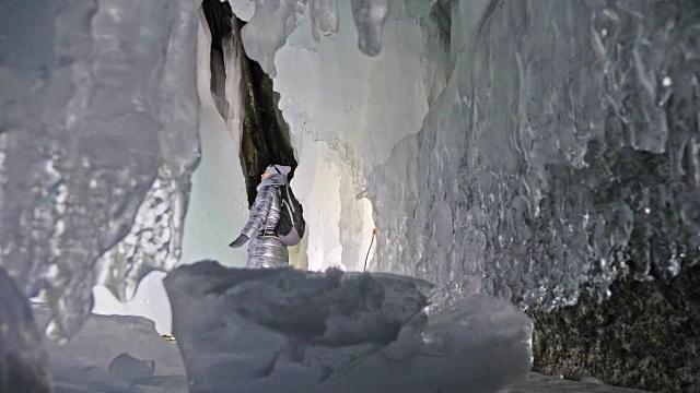 贝加尔湖冰洞上的女游客。冬岛之旅。女孩背包客正在冰洞中行走。游客看着美丽的冰洞。徒步旅行者穿银色夹克，背包，led灯。