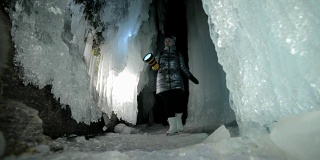贝加尔湖冰洞上的女游客。冬岛之旅。女孩背包客正在冰洞中行走。游客看着美丽的冰洞。徒步旅行者穿银色夹克，背包，led灯。