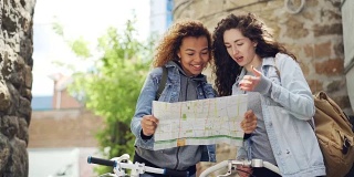 两个游客站在街上，手里拿着自行车，一边看地图一边聊天。迷人的年轻女子和休闲服装与背包，笑着环顾四周。