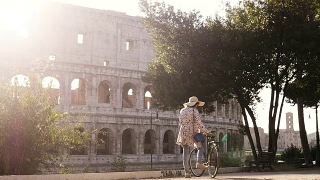 美丽的年轻女子在色彩鲜艳的时装独自行走与自行车在罗马斗牛场前在日落与树快乐迷人的旅游女孩在colle oppio与草帽