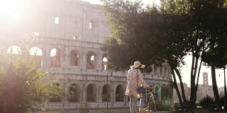 美丽的年轻女子在色彩鲜艳的时装独自行走与自行车在罗马斗牛场前在日落与树快乐迷人的旅游女孩在colle oppio与草帽