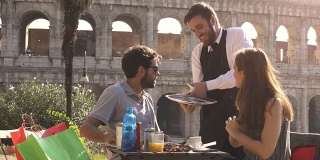 在夕阳西下的罗马斗牛场前，一个酒吧餐厅的桌子上，优雅微笑的服务员正在为一对年轻幸福的夫妇提供早餐和咖啡羊角面包卡布奇诺果汁