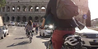 三个年轻的朋友游客在阳光明媚的日子里骑着自行车在罗马市中心的罗马斗牛场周围的道路上慢镜头摄影机汽车稳定摄影机