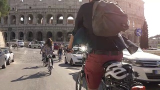三个年轻的朋友游客在阳光明媚的日子里骑着自行车在罗马市中心的罗马斗牛场周围的道路上慢镜头摄影机汽车稳定摄影机视频素材模板下载