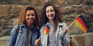 慢动作肖像两个微笑的女孩朋友挥舞着德国国旗，看着相机站在石头墙。友谊、旅游、快乐的人理念。