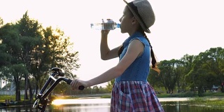 日落时分，一个戴着帽子的小女孩站在河边的公园里，骑着自行车，从一个透明的瓶子里喝水。